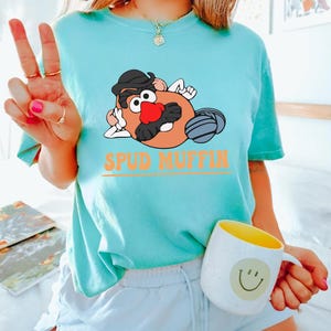 May include: A turquoise blue t-shirt with a cartoon image of Mr. Potato Head wearing a hat and sunglasses. The text "Spud Muffin" is printed in orange below the image.