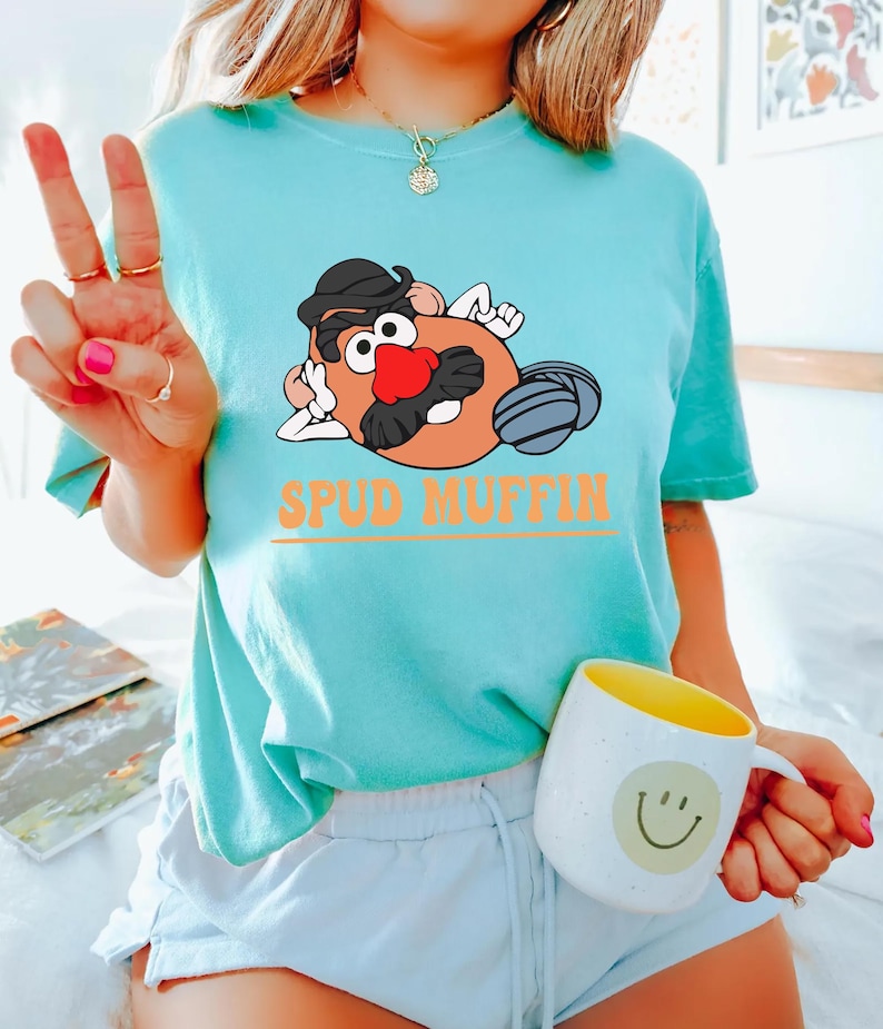 May include: A turquoise blue t-shirt with a cartoon image of Mr. Potato Head wearing a hat and sunglasses. The text "Spud Muffin" is printed in orange below the image.