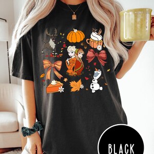 May include: Black t-shirt featuring a colorful fall-themed graphic. The design includes a reindeer, pumpkin, cupcake, bows, and characters from a popular animated film. The word "BLACK" is printed on a black circle at the bottom.