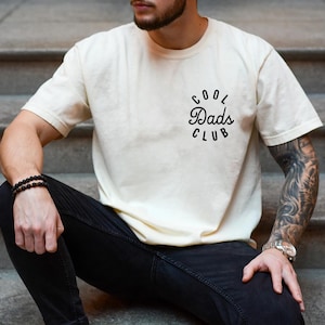 May include: A person wearing a white t-shirt with the text "Cool Dads Club" in black lettering. The person is wearing black jeans and a black beaded bracelet.