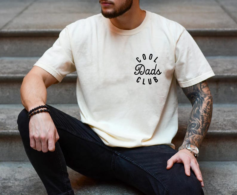 May include: A person wearing a white t-shirt with the text "Cool Dads Club" in black lettering. The person is wearing black jeans and a black beaded bracelet.