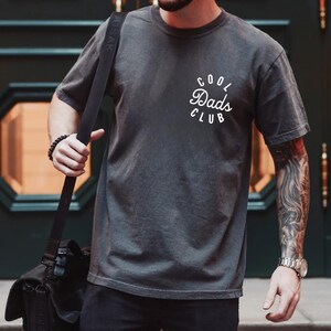 May include: A person wearing a dark gray t-shirt with the text "Cool Dads Club" in white lettering. The person is wearing a black watch on their left wrist and a black beaded bracelet on their right wrist.