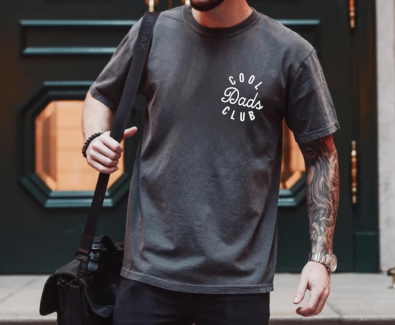 May include: A person wearing a dark gray t-shirt with the text "Cool Dads Club" in white lettering. The person is wearing a black watch on their left wrist and a black beaded bracelet on their right wrist.