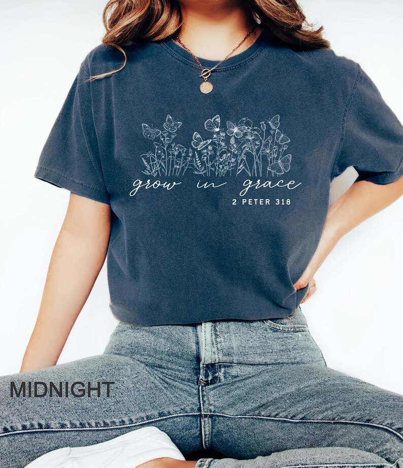 May include: A navy blue t-shirt with a floral and butterfly design. The text "grow in grace" and "2 Peter 3:18" are printed in white. The shirt is paired with blue jeans.