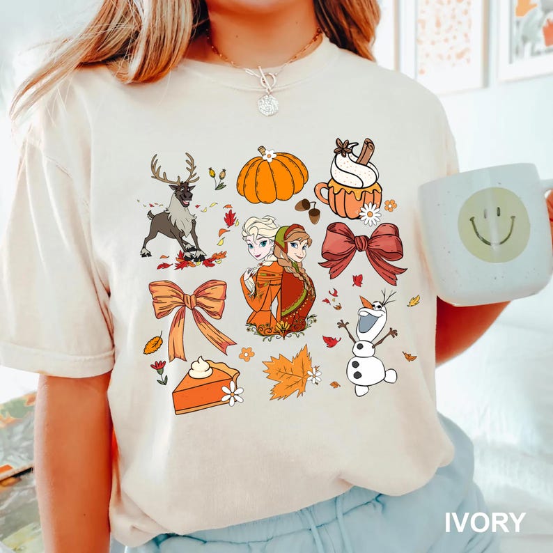May include: Ivory-colored t-shirt featuring a collection of fall-themed illustrations. The design includes a reindeer, pumpkin, bow, pie, and characters from a popular animated film. A white mug with a smiley face is also visible.