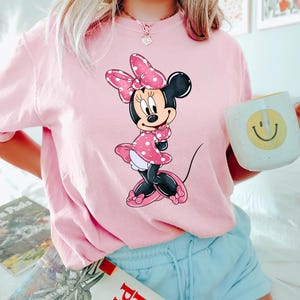 May include: A pink t-shirt featuring a cartoon image of Minnie Mouse wearing a red polka dot dress and a pink bow.