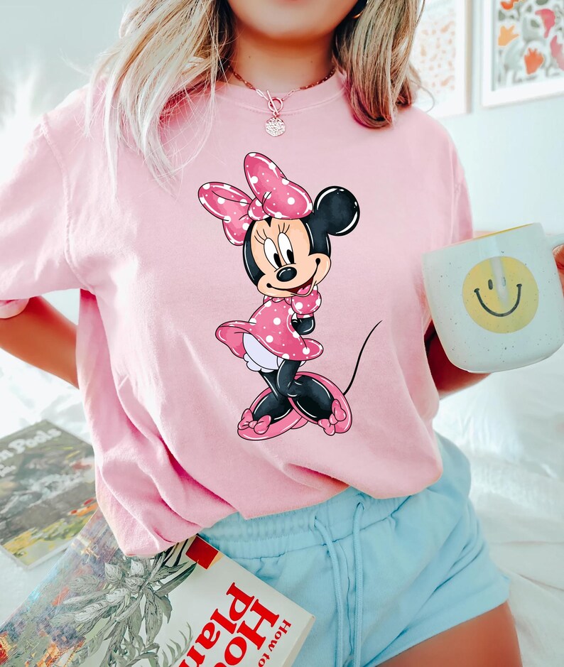 May include: A pink t-shirt featuring a cartoon image of Minnie Mouse wearing a red polka dot dress and a pink bow.