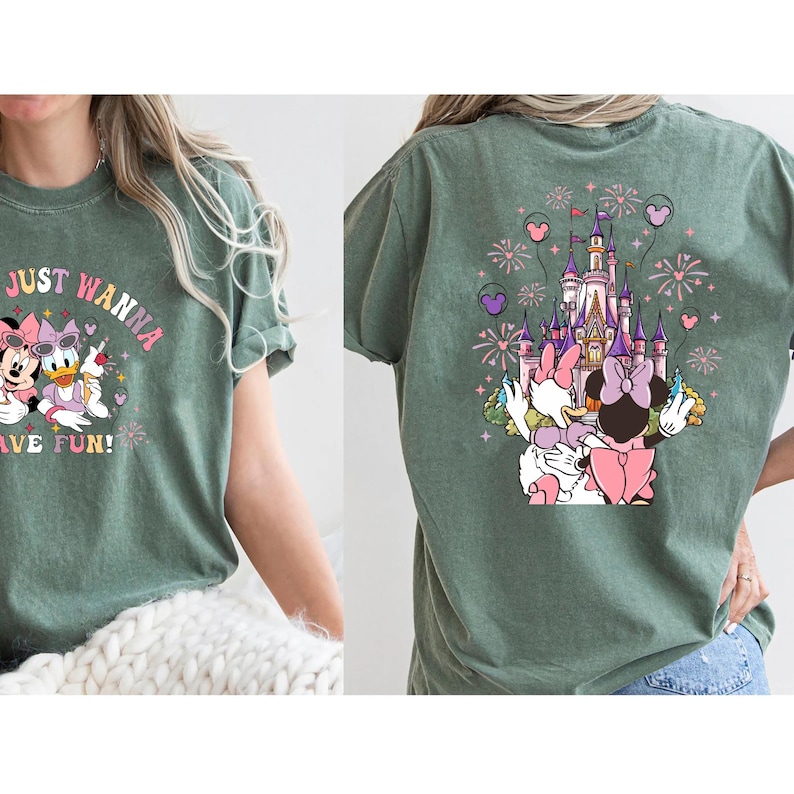 May include: Green t-shirt with a graphic featuring Minnie Mouse, Daisy Duck, and a castle. The front of the shirt says "Just Wanna Have Fun!" in a whimsical font. The back of the shirt shows the castle with fireworks and the characters.