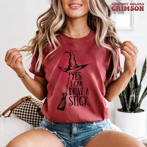 Pu&ograve; includere: T-shirt color cremisi con cappello da strega nero e grafica scopa. Il testo "YES I CAN DRIVE A STICK" &egrave; stampato sotto il cappello. La maglietta &egrave; del marchio Comfort Colors.