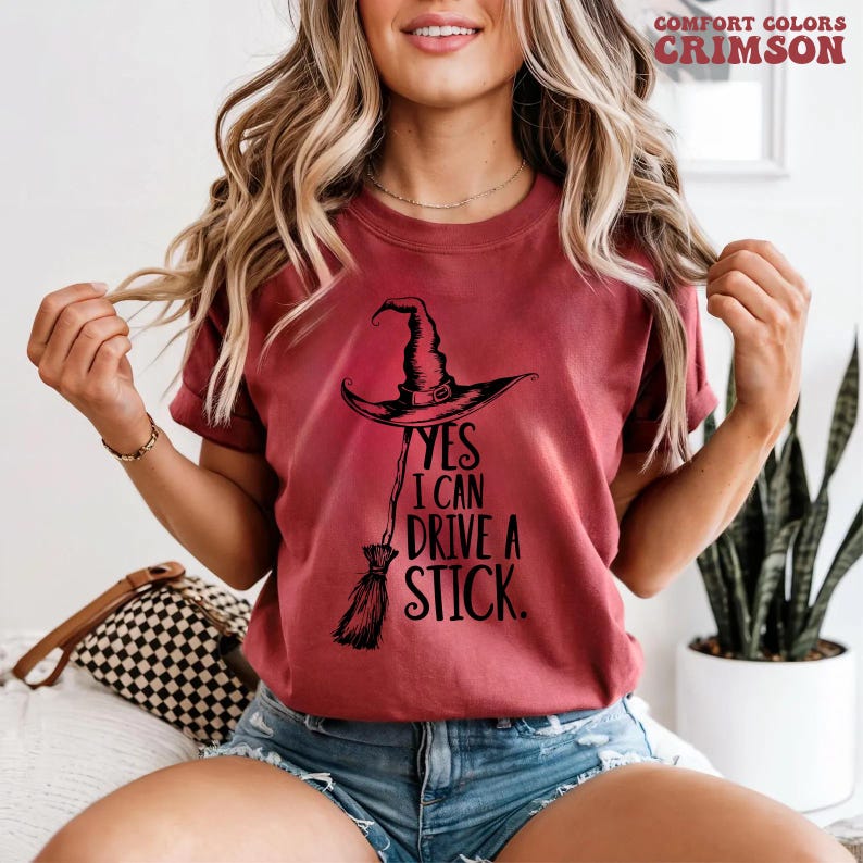 Pu&ograve; includere: T-shirt color cremisi con cappello da strega nero e grafica scopa. Il testo "YES I CAN DRIVE A STICK" &egrave; stampato sotto il cappello. La maglietta &egrave; del marchio Comfort Colors.
