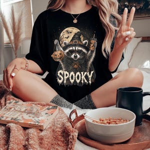 Pu&ograve; includere: Una t-shirt nera con un'illustrazione grafica di un procione che indossa un cappello da strega, tiene in mano una zucca e un bicchiere, con uno sfondo di luna piena e la parola SPOOKY stampata in caratteri bianchi testurizzati.