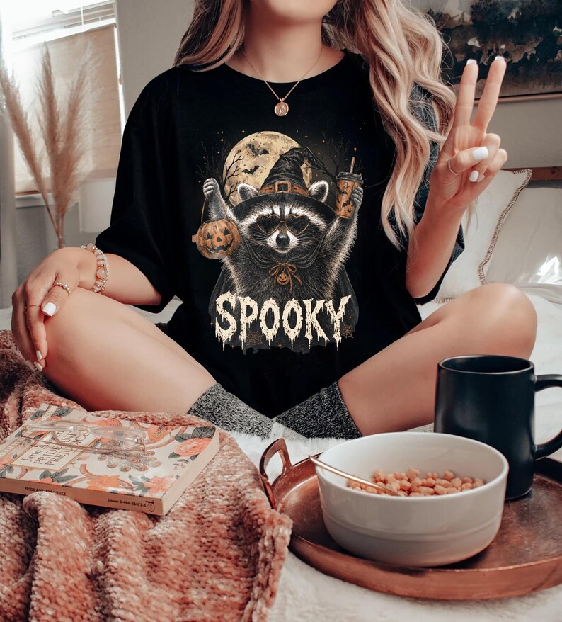 Pu&ograve; includere: Una t-shirt nera con un'illustrazione grafica di un procione che indossa un cappello da strega, tiene in mano una zucca e un bicchiere, con uno sfondo di luna piena e la parola SPOOKY stampata in caratteri bianchi testurizzati.