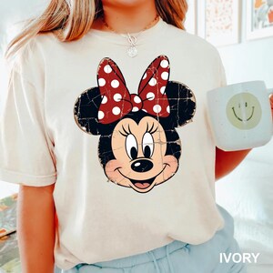 May include: An ivory short-sleeved t-shirt featuring a distressed graphic of Minnie Mouse with a red and white polka dot bow. The character has black ears, a black nose, and a smiling expression. The shirt is paired with a light-coloured mug.