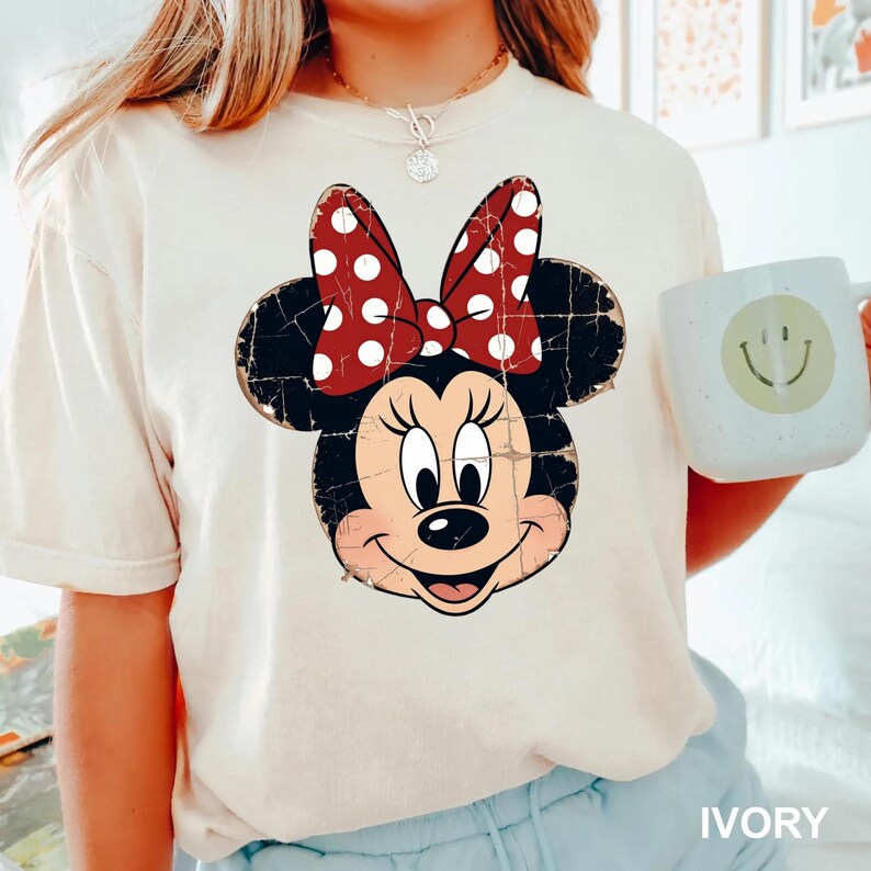 May include: An ivory short-sleeved t-shirt featuring a distressed graphic of Minnie Mouse with a red and white polka dot bow. The character has black ears, a black nose, and a smiling expression. The shirt is paired with a light-coloured mug.