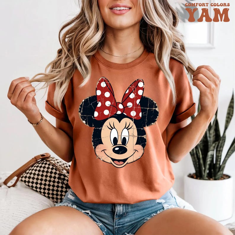 May include: A rust-coloured short-sleeve t-shirt featuring a distressed graphic of Minnie Mouse with a red and white polka dot bow. The shirt has a crew neckline and a relaxed fit. The graphic shows the character's face with a weathered texture.