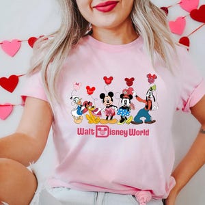 K&ouml;nnte beinhalten: Rosafarbenes T-Shirt mit einem Motiv von Mickey Mouse, Minnie Mouse, Donald Duck, Goofy und Pluto, die Herzen und Ballons halten. Der Text "Walt Disney World" ist unter dem Motiv gedruckt.