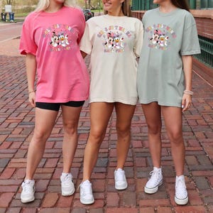 May include: Three women wearing oversized t-shirts in pink, cream, and sage green. Each shirt features a graphic of Minnie Mouse, Daisy Duck, and a text that reads "GIRLS JUST WANNA HAVE FUN!". The women are wearing black shorts and white sneakers.