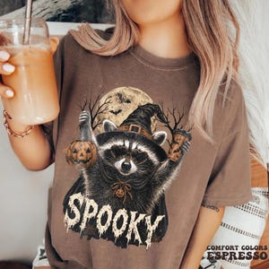 Pu&ograve; includere: Una t-shirt marrone con la grafica di un procione che indossa un cappello da strega e occhiali da sole a forma di cuore. Il procione tiene in mano un secchiello a forma di zucca e un bicchiere. La parola SPOOKY &egrave; stampata in bianco.