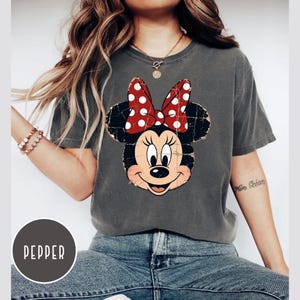 May include: A charcoal grey short-sleeve t-shirt featuring a distressed graphic of Minnie Mouse with a red and white polka dot bow. The shirt has a crew neckline and a relaxed fit. A circular label in the corner displays the text PEPPER.