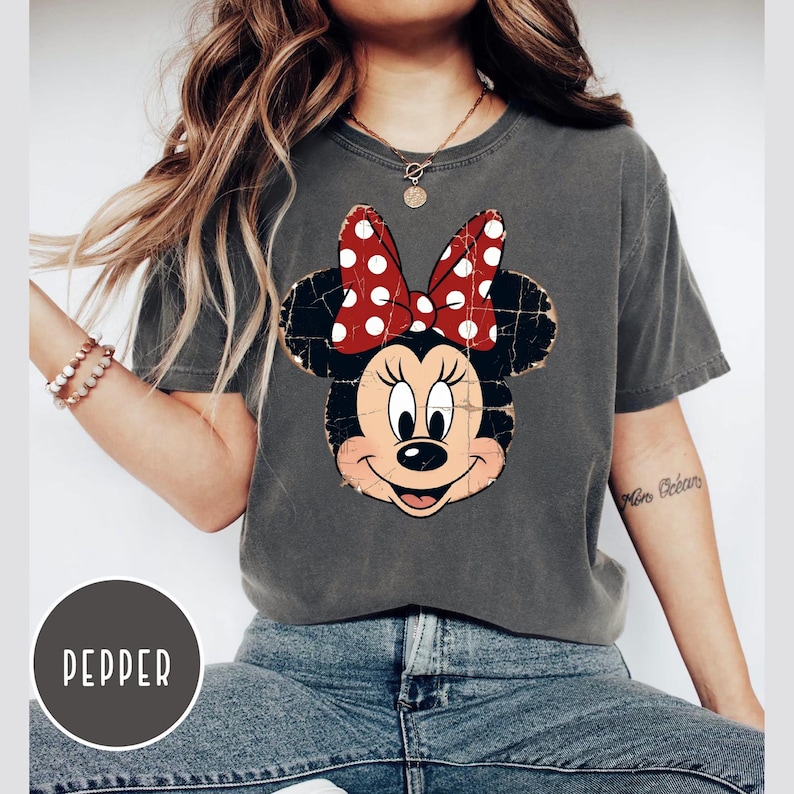 May include: A charcoal grey short-sleeve t-shirt featuring a distressed graphic of Minnie Mouse with a red and white polka dot bow. The shirt has a crew neckline and a relaxed fit. A circular label in the corner displays the text PEPPER.