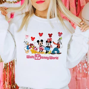 K&ouml;nnte beinhalten: Wei&szlig;er Pullover mit einem Grafikdruck von Mickey Mouse, Minnie Mouse, Donald Duck, Goofy und Pluto, die rote und rosa Herzen halten. Der Text "Walt Disney World" ist unter den Figuren gedruckt.