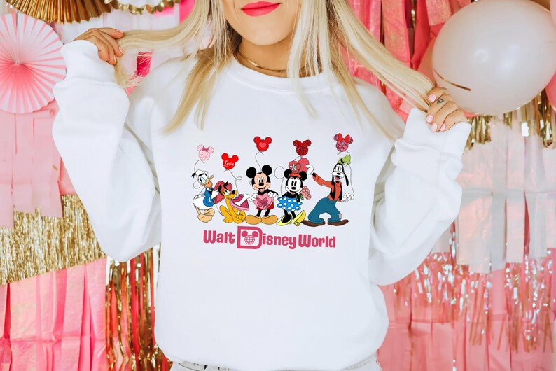 K&ouml;nnte beinhalten: Wei&szlig;er Pullover mit einem Grafikdruck von Mickey Mouse, Minnie Mouse, Donald Duck, Goofy und Pluto, die rote und rosa Herzen halten. Der Text "Walt Disney World" ist unter den Figuren gedruckt.