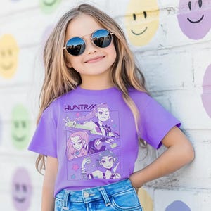 Peut inclure: Une jeune personne porte un t-shirt violet avec le texte "HUNTRA" et des illustrations de style anime. La personne porte &eacute;galement des lunettes de soleil rondes et un jean bleu. Le fond est un mur de briques blanches avec des motifs de visages souriants.