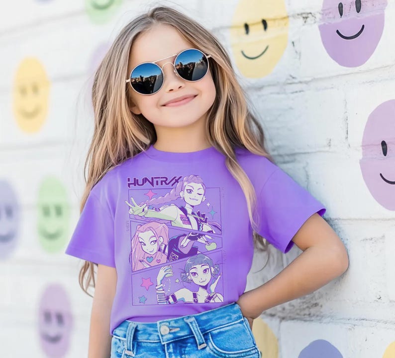 Peut inclure: Une jeune personne porte un t-shirt violet avec le texte "HUNTRA" et des illustrations de style anime. La personne porte &eacute;galement des lunettes de soleil rondes et un jean bleu. Le fond est un mur de briques blanches avec des motifs de visages souriants.