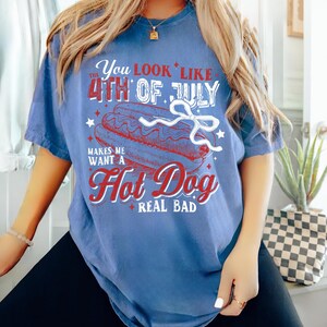 Pode incluir: Uma camiseta azul com um gr&aacute;fico branco que diz "You look like the 4th of July makes me want a hot dog real bad". O gr&aacute;fico inclui um cachorro-quente vermelho, branco e azul com mostarda e picles.