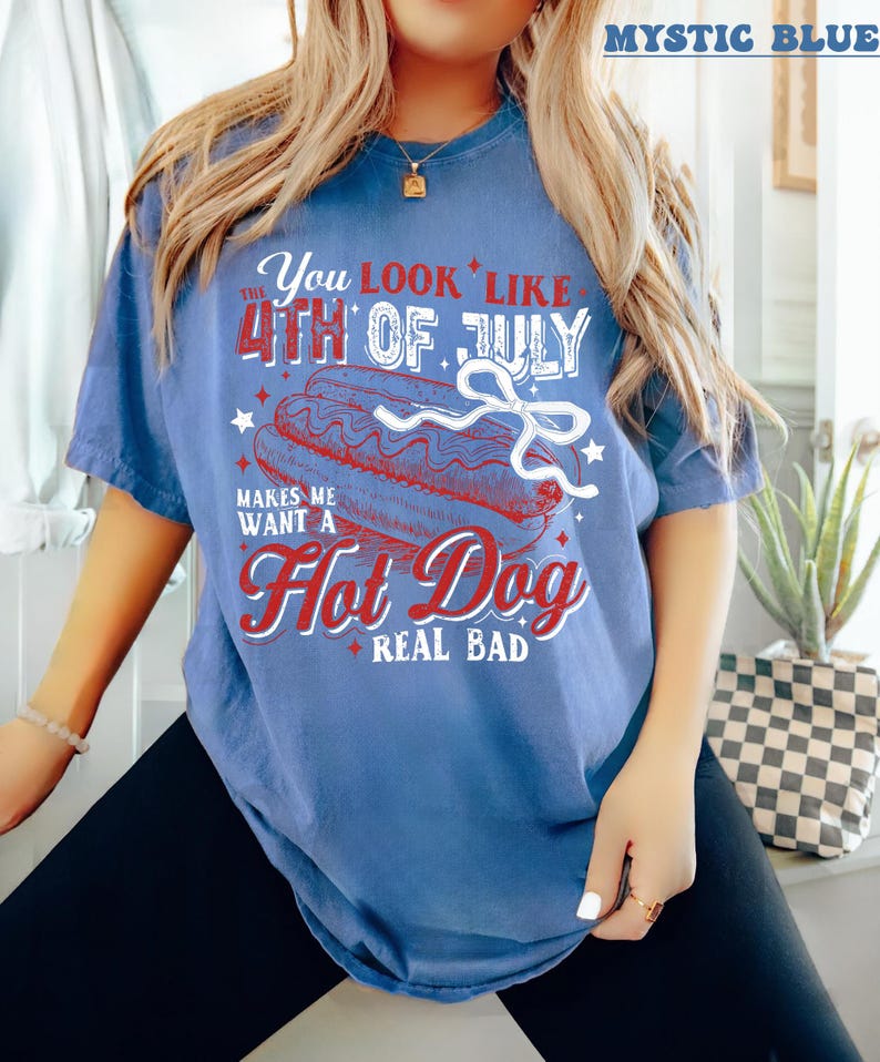 Pode incluir: Uma camiseta azul com um gr&aacute;fico branco que diz "You look like the 4th of July makes me want a hot dog real bad". O gr&aacute;fico inclui um cachorro-quente vermelho, branco e azul com mostarda e picles.