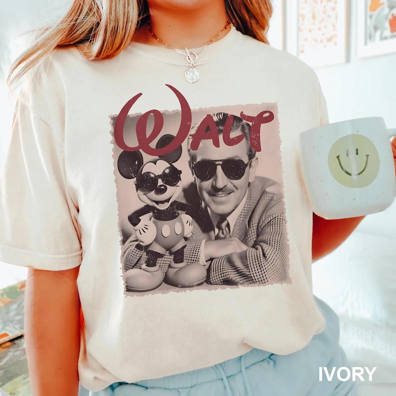 Op de afbeelding: Een ivoorkleurig t-shirt met korte mouwen en een grafische print van een man met een zonnebril naast een Mickey Mouse-figuur met een zonnebril. Het woord Walt staat in grote kastanjebruine letters boven de figuren. Een lichte mok is zichtbaar.