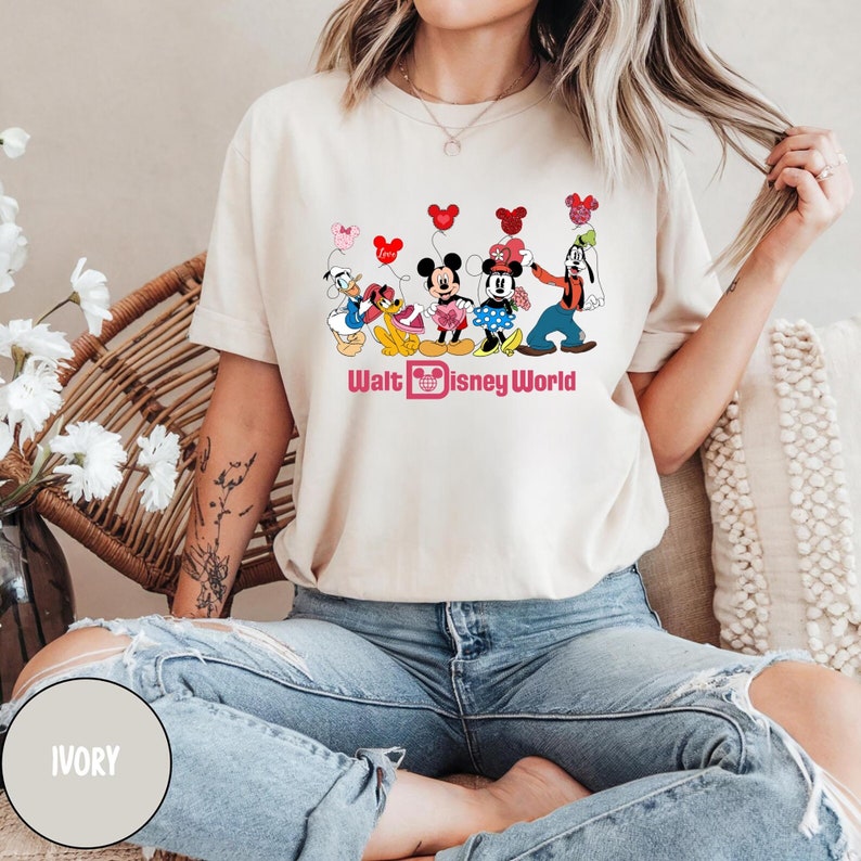 K&ouml;nnte beinhalten: Ein wei&szlig;es T-Shirt mit einem Aufdruck von Mickey Mouse, Minnie Mouse, Donald Duck, Goofy und Pluto, die rote, rosa und wei&szlig;e Ballons halten. Der Text "Walt Disney World" ist in Pink unter dem Aufdruck gedruckt.