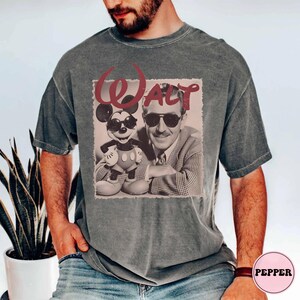 Op de afbeelding: Een antracietgrijs T-shirt met korte mouwen en een grafische print van Walt Disney en Mickey Mouse, beiden met een zonnebril op. De tekst "WALT" is in een groot, gestileerd rood lettertype boven de figuren op een lichte achtergrond gedrukt.
