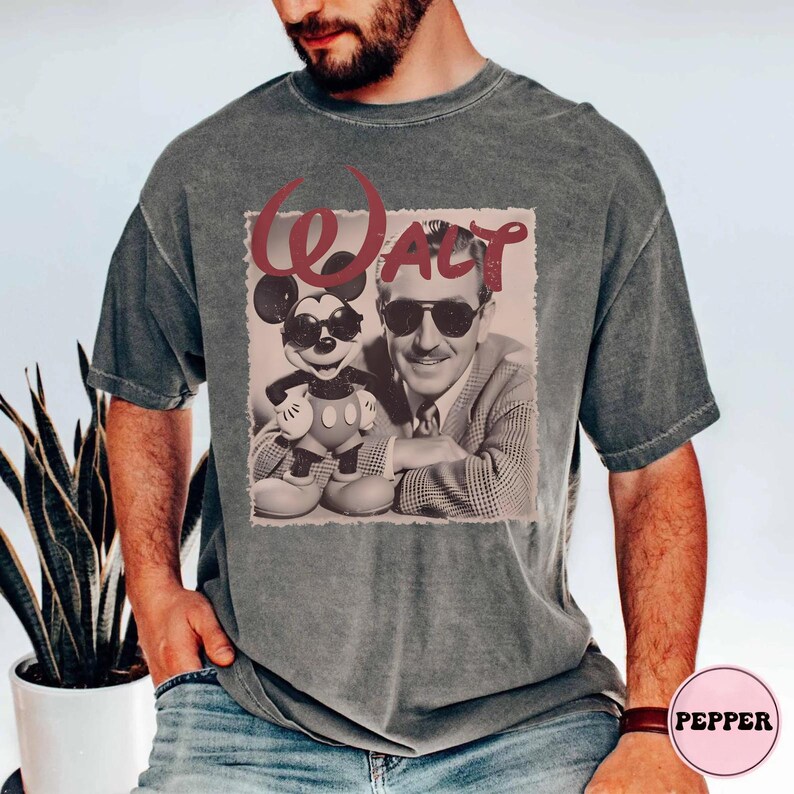 Op de afbeelding: Een antracietgrijs T-shirt met korte mouwen en een grafische print van Walt Disney en Mickey Mouse, beiden met een zonnebril op. De tekst "WALT" is in een groot, gestileerd rood lettertype boven de figuren op een lichte achtergrond gedrukt.