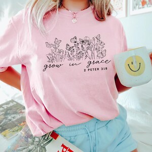 May include: A light pink t-shirt with a black floral and butterfly design, and the words "grow in grace" and "2 Peter 3:18". A white mug with a yellow smiley face is held in the hand.