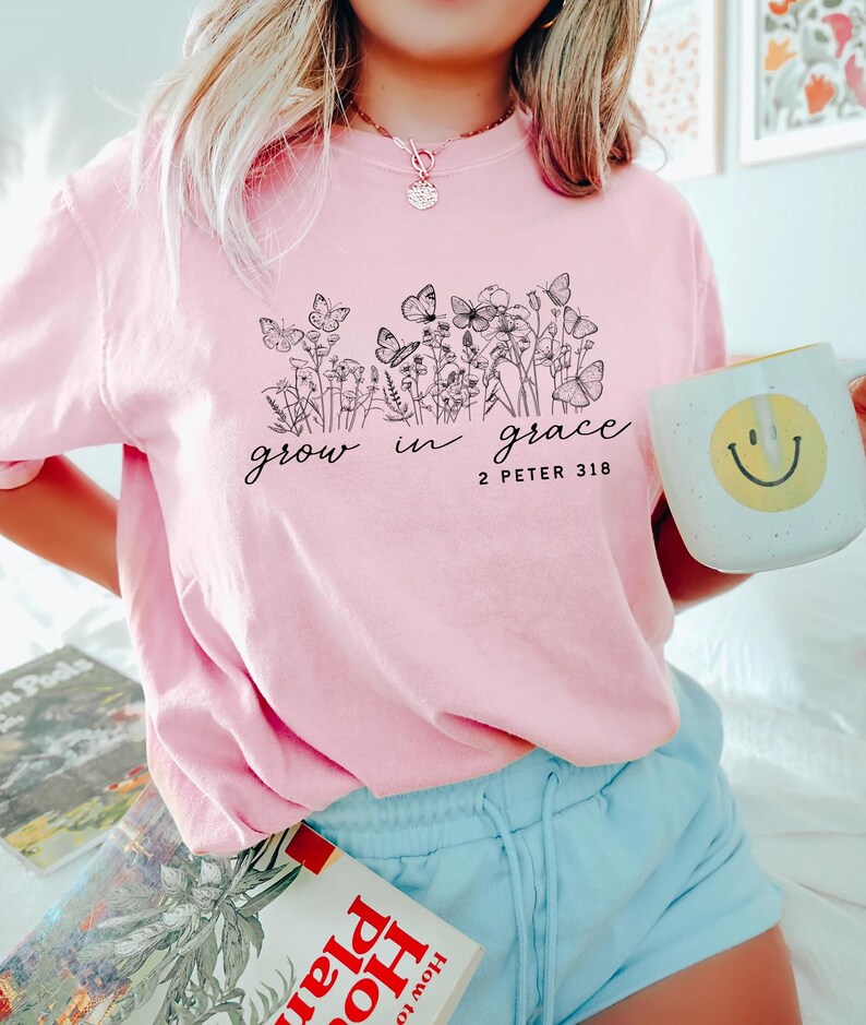 May include: A light pink t-shirt with a black floral and butterfly design, and the words "grow in grace" and "2 Peter 3:18". A white mug with a yellow smiley face is held in the hand.