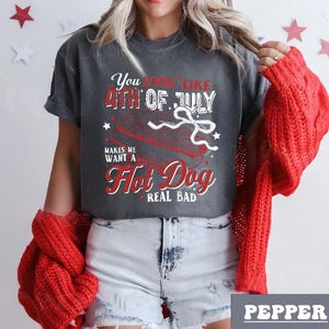 Pode incluir: Uma camiseta cinza com um gr&aacute;fico vermelho e branco que diz "You look like the 4th of July makes me want a Hot Dog real bad". O gr&aacute;fico apresenta um cachorro-quente com uma fita vermelha, branca e azul.