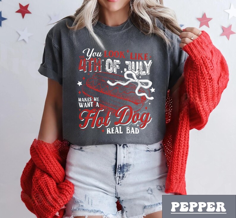 Pode incluir: Uma camiseta cinza com um gr&aacute;fico vermelho e branco que diz "You look like the 4th of July makes me want a Hot Dog real bad". O gr&aacute;fico apresenta um cachorro-quente com uma fita vermelha, branca e azul.