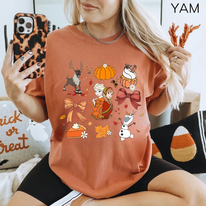 May include: Rust-colored t-shirt featuring a collection of fall-themed illustrations. The design includes a reindeer, pumpkins, pie, bows, autumn leaves, and characters from the movie Frozen. The shirt is a comfortable, casual style.