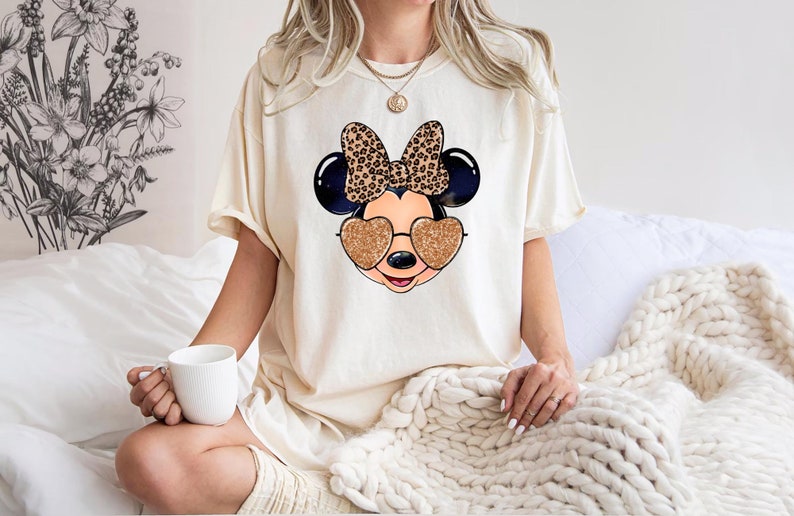 以下が含まれることがあります： レオパード柄のリボンとハート型のサングラスをかけたマウスの漫画の画像がプリントされたクリーム色のTシャツ。