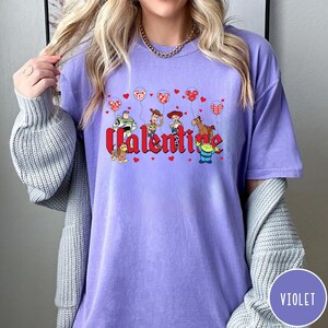 May include: Purple t-shirt with a graphic of Toy Story characters holding red heart balloons and the word "Valentine".