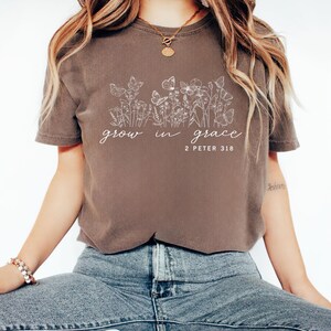 May include: A brown t-shirt with the words "grow in grace" and the Bible verse "2 Peter 3:18" in white script. The shirt features a floral and butterfly design. The model is wearing blue jeans.