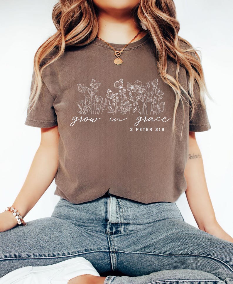 May include: A brown t-shirt with the words "grow in grace" and the Bible verse "2 Peter 3:18" in white script. The shirt features a floral and butterfly design. The model is wearing blue jeans.
