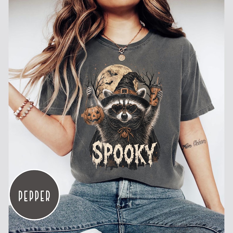 Pu&ograve; includere: Una t-shirt a maniche corte grigio antracite con la grafica di un procione che indossa un cappello da strega e occhiali da sole. Il procione tiene in mano una zucca e un bicchiere di caff&egrave; sotto una luna piena. La scritta SPOOKY &egrave; in bianco.