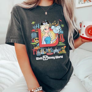 Może przedstawiać: Szary t-shirt z kolorowym nadrukiem graficznym przedstawiającym postacie i atrakcje Disneya. Grafika zawiera rysunkową postać Elsy z Krainy Lodu, Myszki Miki i napis "Walt Disney World".