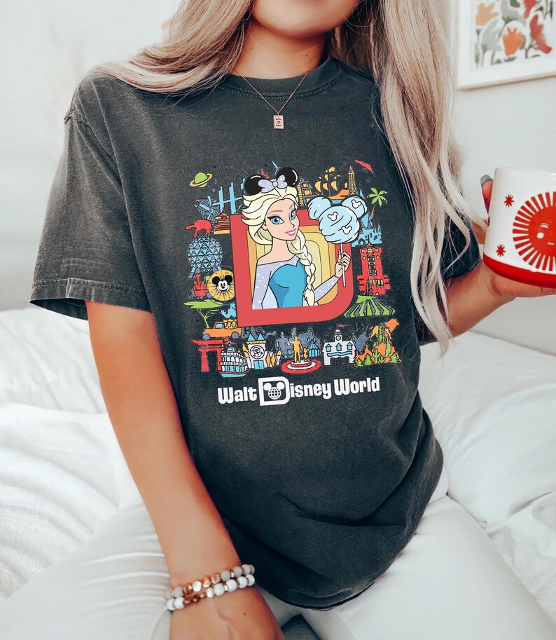 Może przedstawiać: Szary t-shirt z kolorowym nadrukiem graficznym przedstawiającym postacie i atrakcje Disneya. Grafika zawiera rysunkową postać Elsy z Krainy Lodu, Myszki Miki i napis "Walt Disney World".