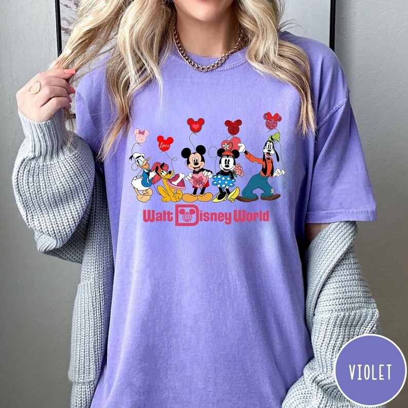 K&ouml;nnte beinhalten: Ein lila T-Shirt mit einem Aufdruck von Mickey Mouse, Minnie Mouse, Donald Duck, Goofy und Pluto mit Herzen und Ballons. Der Text "Walt Disney World" ist unter den Figuren gedruckt.
