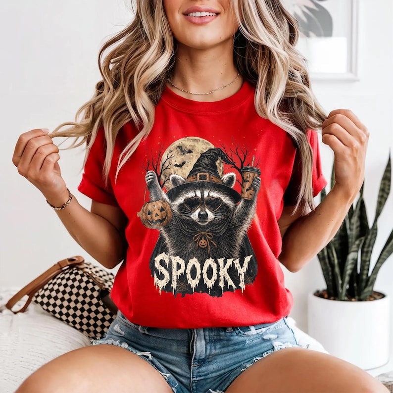 Pu&ograve; includere: Una t-shirt rosso brillante con la grafica di un procione che indossa un cappello da strega e occhiali. Il procione tiene in mano una zucca e un bicchiere. Dietro ci sono una luna piena e alberi spogli. La parola SPOOKY &egrave; stampata in bianco.