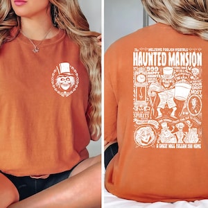 May include: A burnt orange t-shirt with a white graphic print of the Haunted Mansion attraction at Disney World. The graphic includes the text "Welcome Foolish Mortals Haunted Mansion 999 Happy Haunts Your Ghost Host Spirits with Madame Leota Coming Soon Get Out to Sooner A Ghost Will Follow You Home".