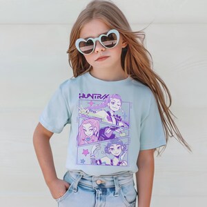Peut inclure: Un t-shirt bleu clair avec un graphisme de trois personnages de style anime aux accents violets et roses. Le mot "HUNTRA" est imprim&eacute; au-dessus du graphisme en violet. Le t-shirt est port&eacute; par une jeune personne portant des lunettes de soleil en forme de c&oelig;ur.